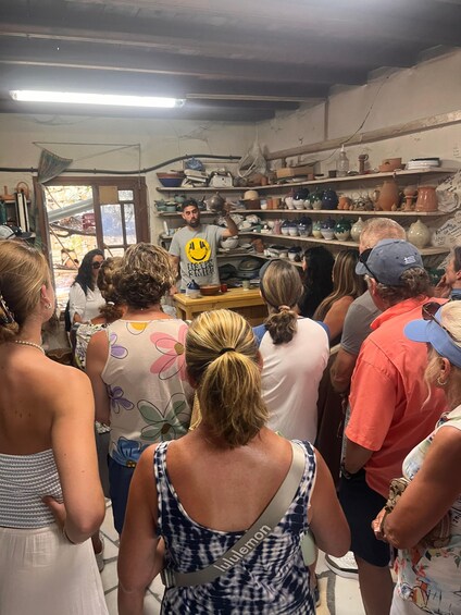 A group of people in a pottery workshop with shelves of pottery and a person demonstrating pottery techniques. 3