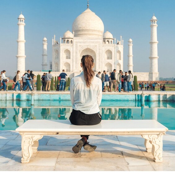 A person sitting on a bench facing the Taj Mahal, with a pool and visitors in the foreground. 3