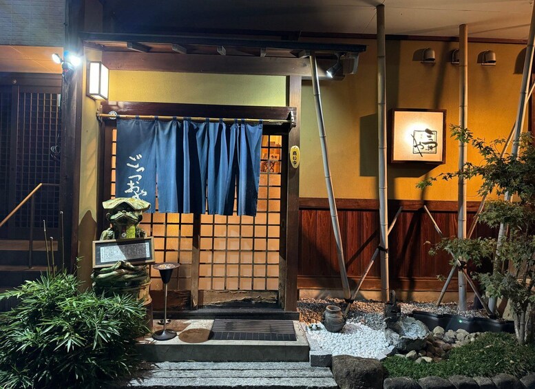 A traditional Japanese entrance with a wooden gate, blue curtains, and a small garden with rocks and plants. 2