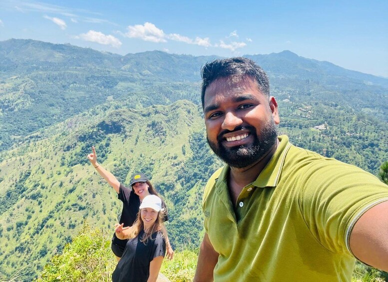 A person in a green shirt taking a selfie with two others in the background, overlooking a mountainous landscape. 3