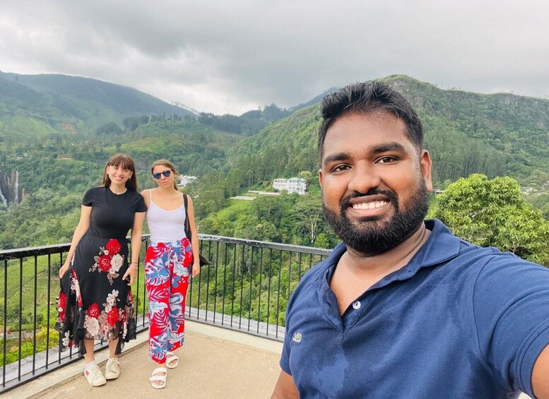 A person taking a selfie with a scenic view of a lush green valley and a waterfall in the background. 1