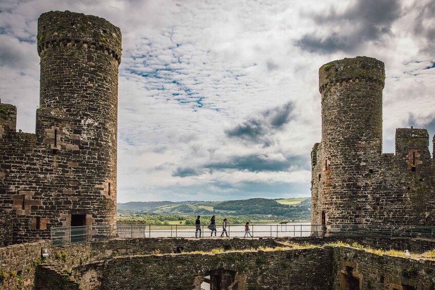 A historic castle with two prominent towers, a river in the background, and a group of people walking along a path. 4