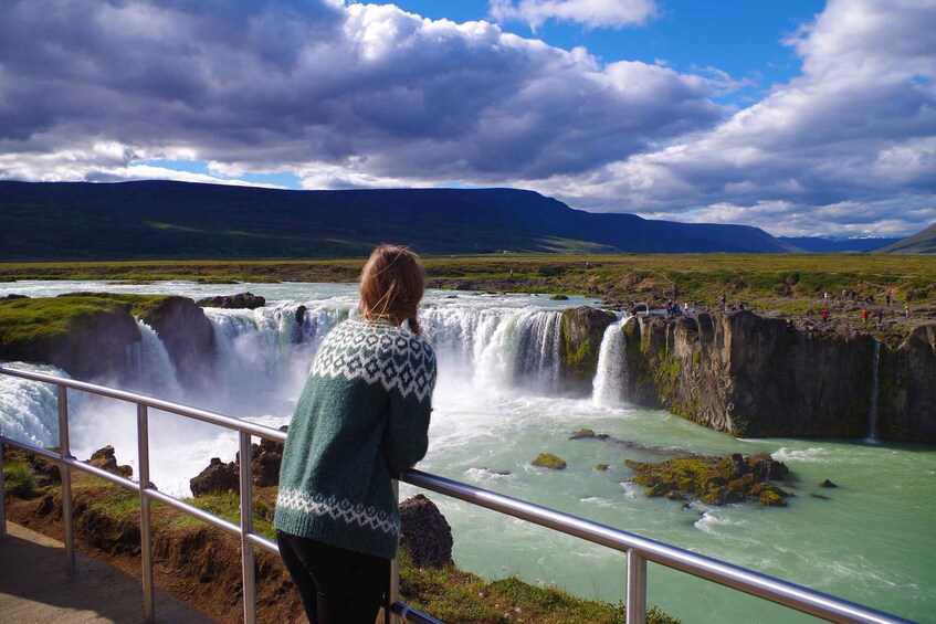 A waterfall with multiple cascades, a person observing from a railing, and a mountainous landscape in the background. 2