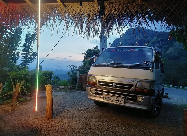 A Toyota Hiace van parked under a thatched roof structure with a mountainous backdrop. 1
