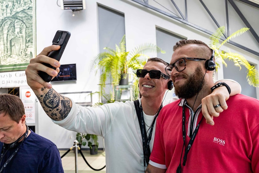 Two people taking a selfie with a smartphone. One person is wearing a white shirt and has a tattoo on their arm. The other person is wearing a red shirt with the word "BOSS" on it. There are plants and a sign in the background. 1