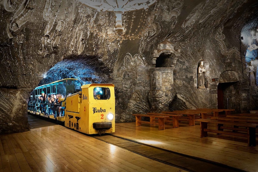 A yellow tram labeled 'Kuba 2' in a cave-like interior with wooden benches and stone carvings. 1