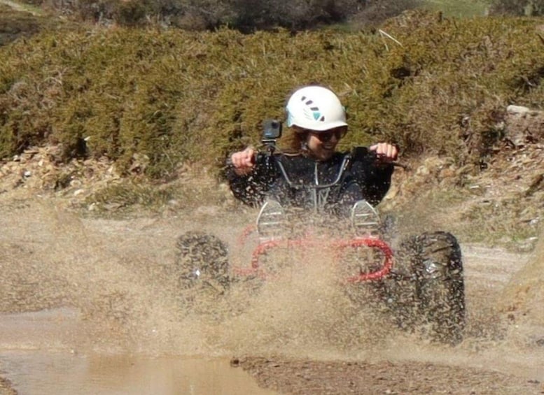 A person in a white helmet and black wetsuit riding a red ATV through a muddy area. 1