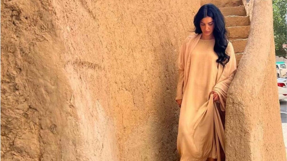 A person in a beige dress standing next to a large stone structure. 1
