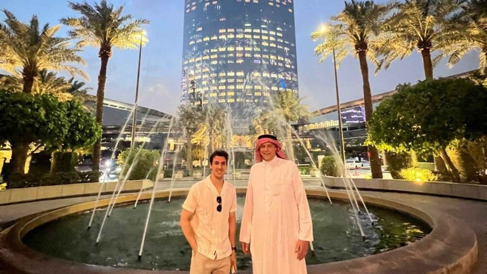 Two men standing in front of a fountain with palm trees and a tall building in the background. 4
