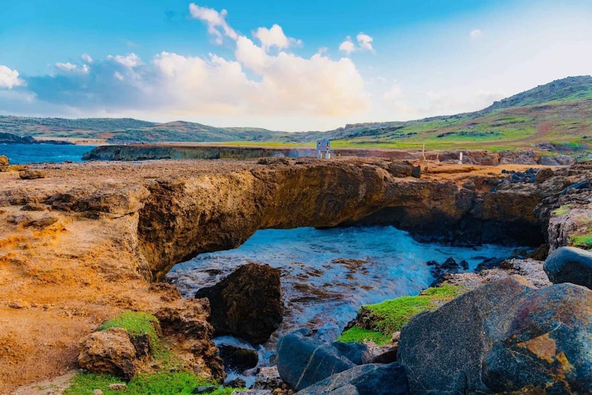 A coastal landscape with a rocky arch formation, a grassy hill in the background, and a clear sky. 1