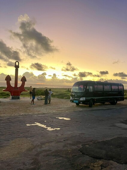 A sunset with a red anchor sculpture, a green van, and people walking on a dirt path. 4