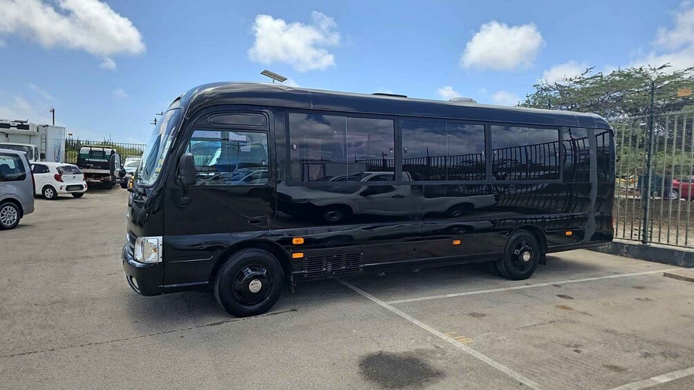 A black bus parked in a lot with other vehicles, a clear sky, and a fence in the background. 3