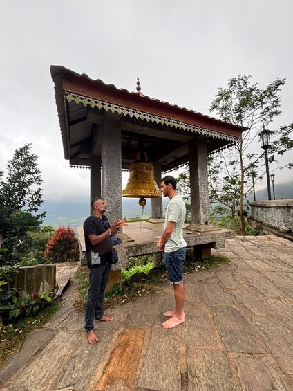 Two people standing on a stone platform with a bell in front of a temple structure. 1