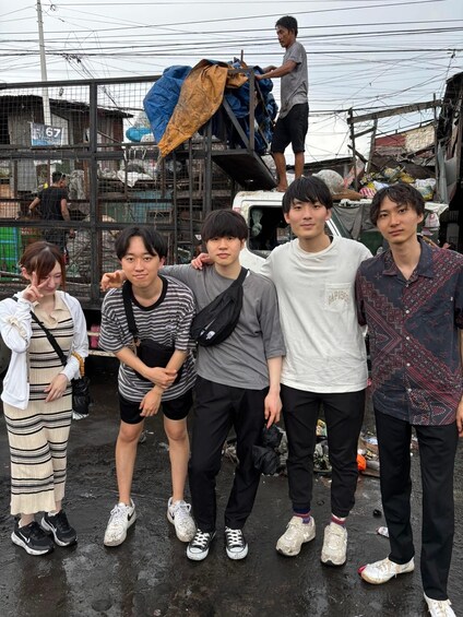 A group of people standing in an outdoor setting with a makeshift structure and a vehicle in the background. 4