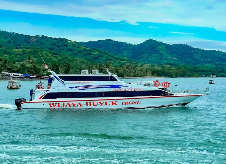 Ein weißes Boot mit roten und blauen Streifen, genannt 'WlJaya Buyuk Cruise', auf ruhigem Wasser vor grünen Hügeln. 2