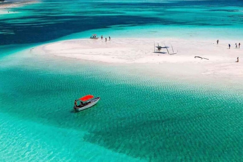 An aerial view of a turquoise lagoon with a small island, a boat, and a few people on the shore. 4
