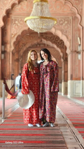 Two people in patterned pajamas standing in a hallway with ornate arches and a chandelier. 1