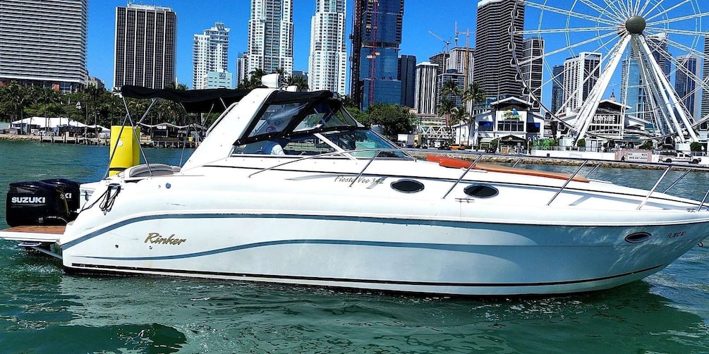 A white boat named 'Rink' docked near a city waterfront with tall buildings and a Ferris wheel in the background. 2