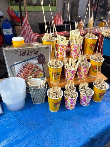 A food stall with skewered snacks in colorful cups, a sign for 'Fish Cake' priced at P50, and a blue tarp covering the table. 3