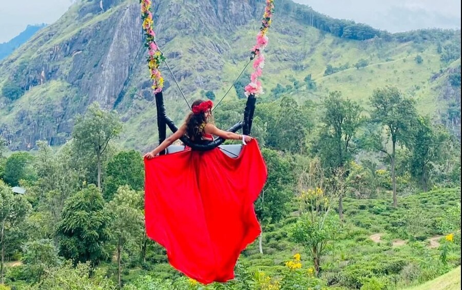 A person on a swing with a red dress, surrounded by greenery and mountains. 1