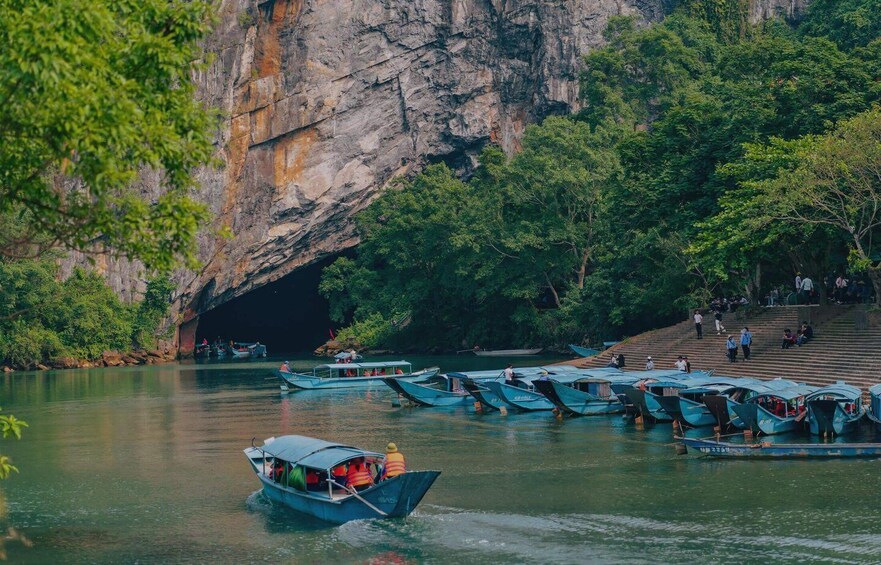 A river with boats docked along the shore, a cave entrance, and people walking on a staircase. 3