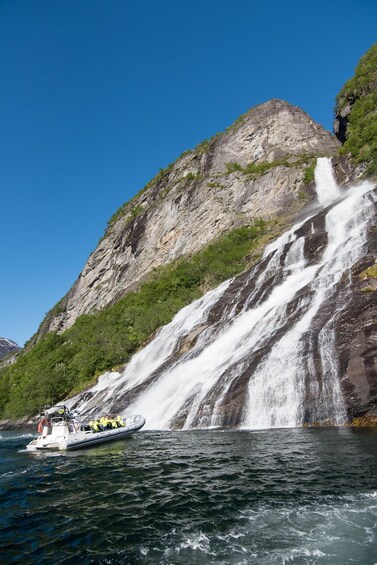A boat near a large waterfall with a rocky cliff and greenery. 1