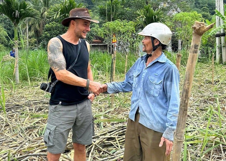 Two people shaking hands in a rural setting with vegetation and a wooden structure in the background. 4