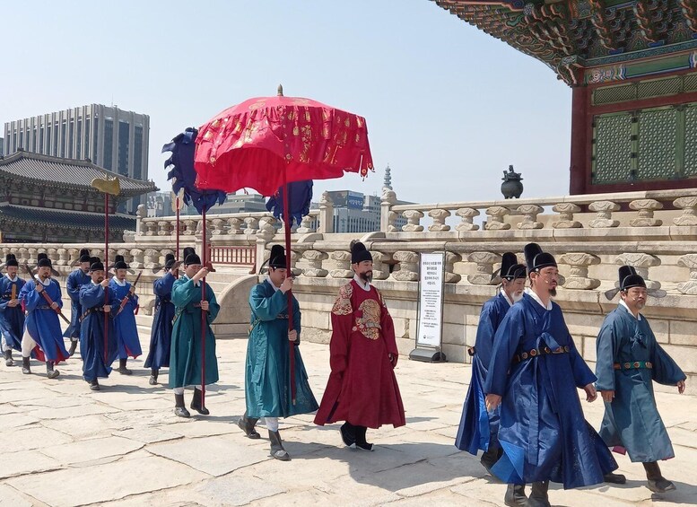 A group of people in traditional attire walking in a line, holding red umbrellas, with a historic building in the background. 1