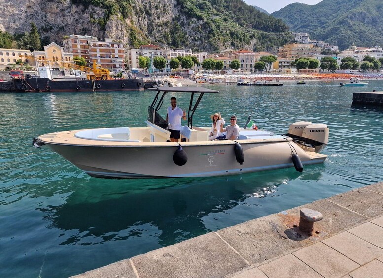 A boat with three people on it, docked near a waterfront with buildings and a rocky hillside in the background. 5
