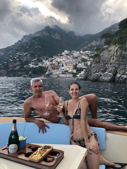 Two people on a boat with a bottle of champagne, snacks, and a scenic view of a coastal village with buildings on cliffs. 3