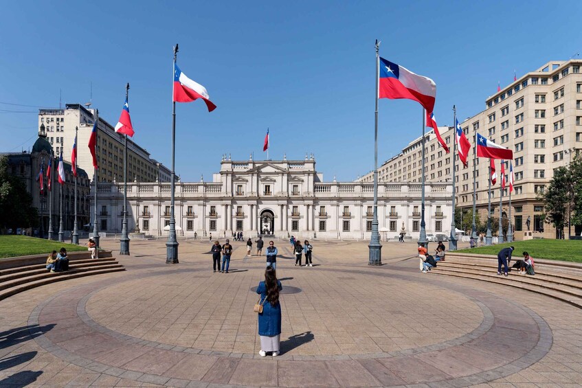 A plaza with a central building, surrounded by flags, people walking, and benches. 1