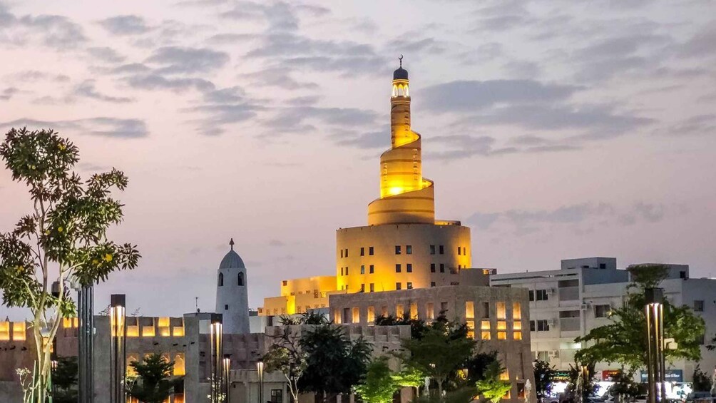 A large, illuminated minaret with a spiral design, surrounded by modern buildings and trees. 5