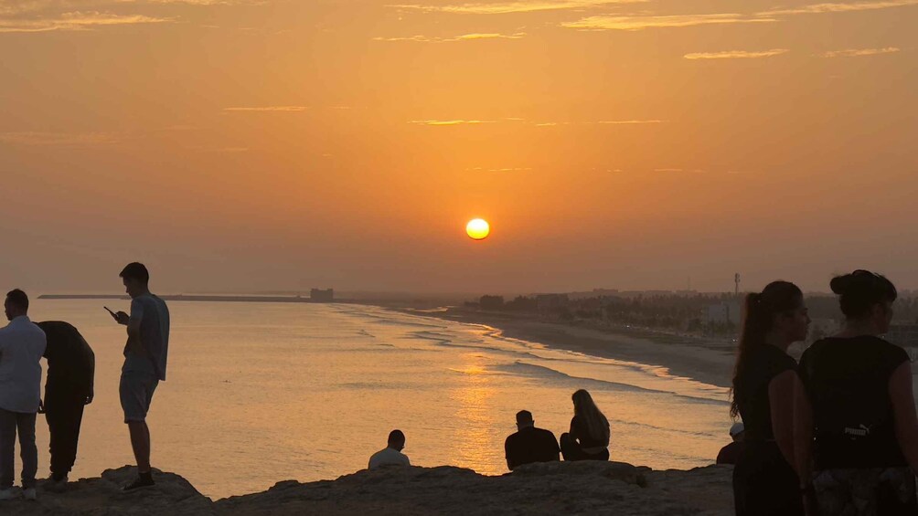 A sunset over a beach with people standing and sitting on the sand. 1