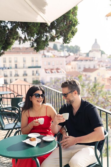 Two people sitting at a table outdoors, holding cups, with buildings in the background. 5