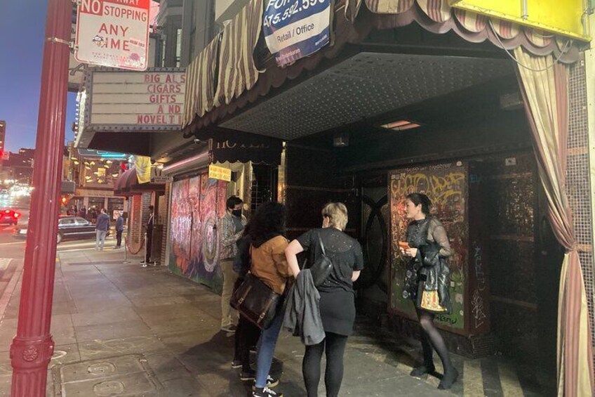 Une scène de rue où l’on voit des personnes se tenir devant un bâtiment orné de graffitis et sur lequel est apposée une enseigne indiquant : « BIG ALS CIGARS GIFTS AND NOVELTY ». 4