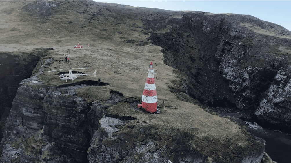 A coastal landscape with a lighthouse, rocky cliffs, and a helicopter on the ground. 1