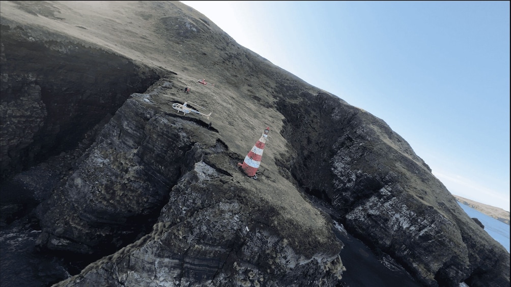 A rocky cliff with a red and white striped pole, a helicopter, and a person standing on the cliff edge. 2