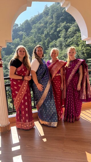 Four women in traditional attire standing in an outdoor setting with a wooden deck and a mountainous backdrop. 2