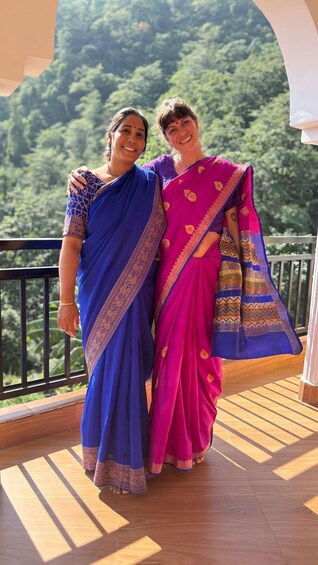 Two women in traditional attire on a wooden balcony with a mountainous landscape in the background. 3