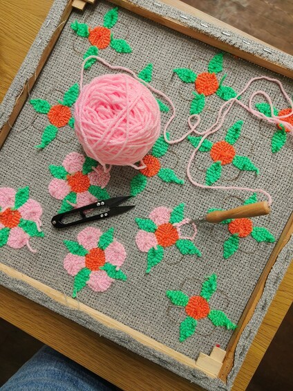 Haakwerk op houten frame met roze en groene wolbolletjes, scharen en een haaknaald. 2