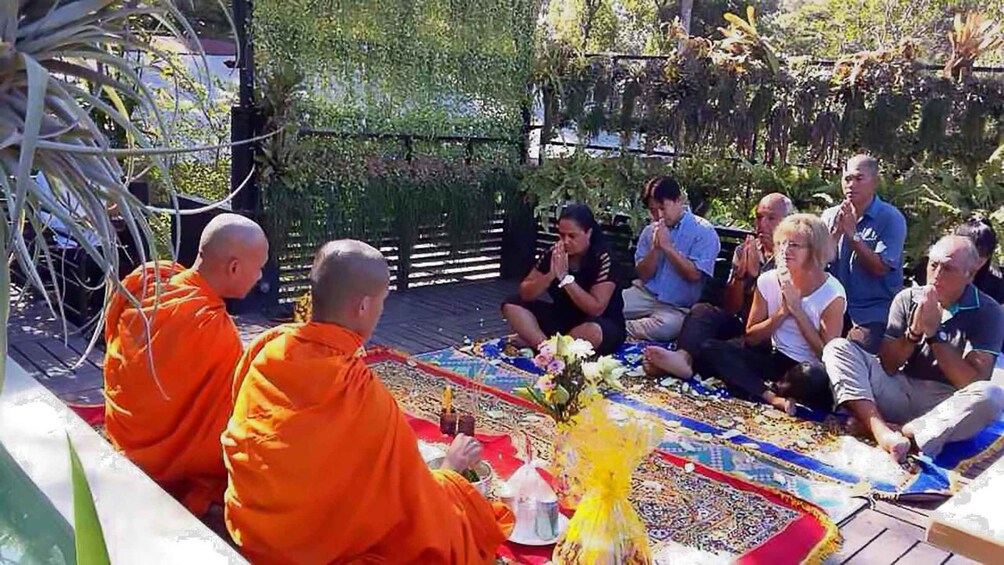 Monks in orange robes sitting on a colorful mat outdoors, surrounded by people. 4