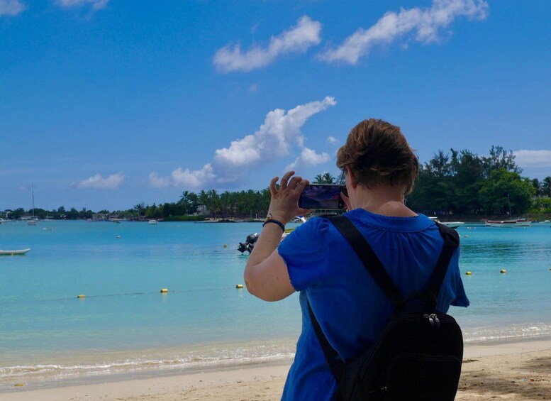 A person taking a photo of a beach with clear blue water, a sandy shore, and a line of trees in the background. 2