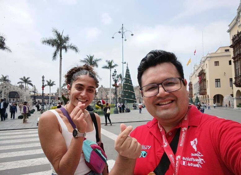 Two people taking a selfie with a historic plaza, palm trees, and a Christmas tree in the background. 1