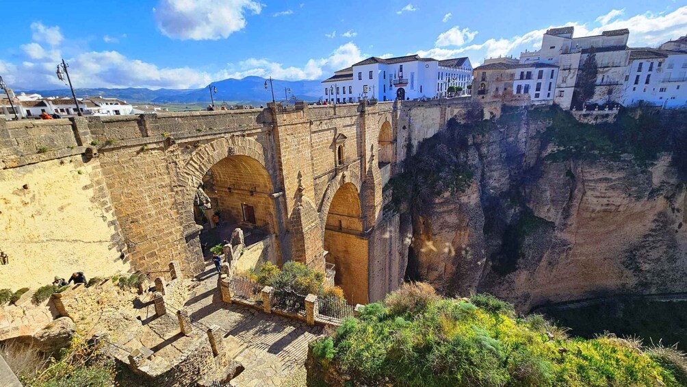 A historic stone bridge with arches, a steep cliff, and white buildings in the background. 1