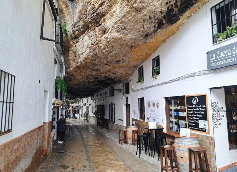 A narrow street with a rock overhang, white buildings, and outdoor seating for cafes. 4