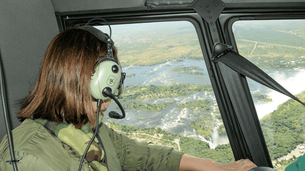 A person in a green flight suit inside a helicopter cockpit, looking out at a landscape with water bodies and greenery. 1