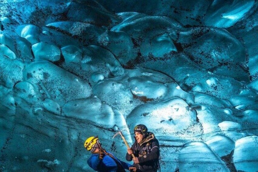 A person in a blue jacket and helmet, holding a hammer, in a crevasse with ice walls. 4
