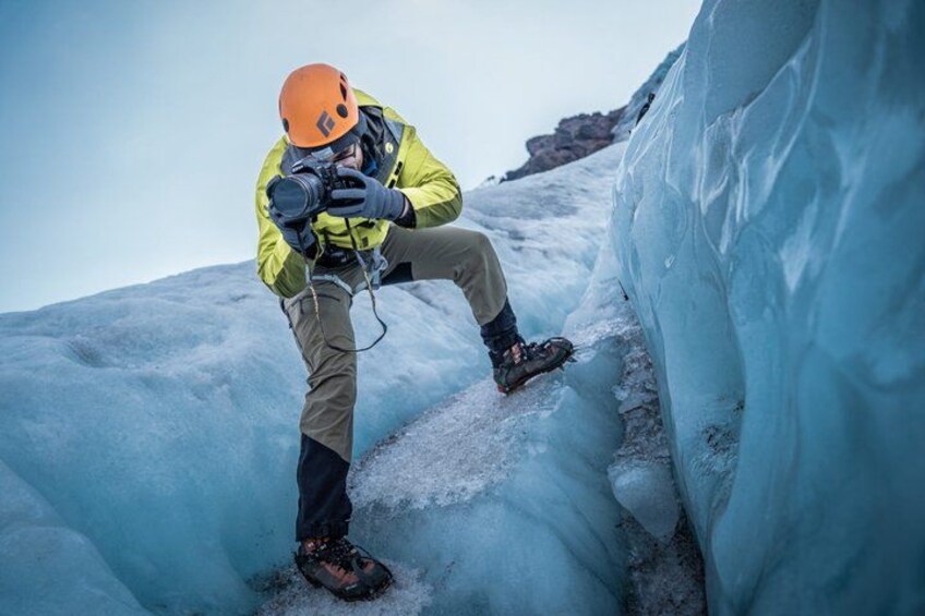 A person in a yellow jacket and orange helmet taking a photograph in a snowy crevasse. 2