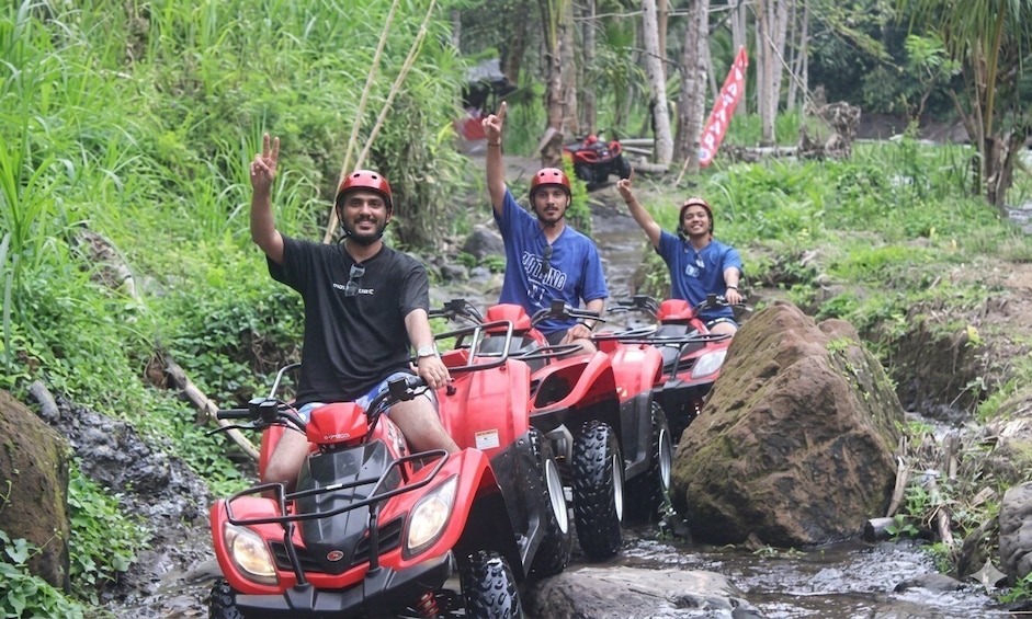 Three people on red ATVs navigating a rocky stream in a forested area. 1