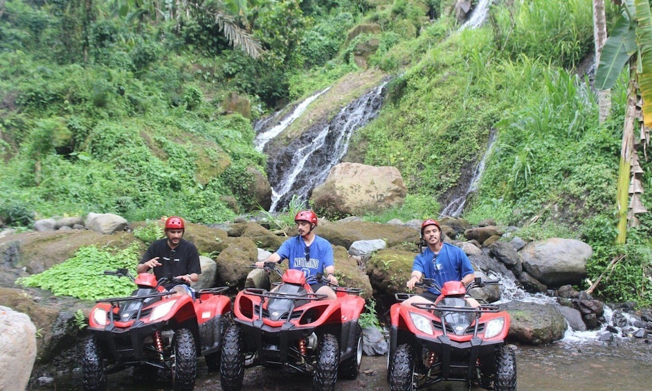 Three people on red ATVs in a lush, green environment with a waterfall in the background. 2